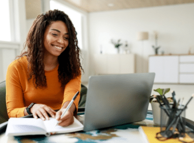 A woman sits at a table with a laptop and pen, focused on creating a budget plan.