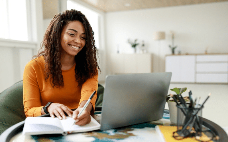 A woman sits at a table with a laptop and pen, focused on creating a budget plan.