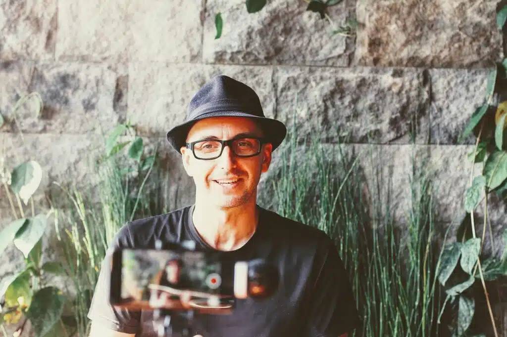 A man wearing a sun hat and glasses is recording a video outdoors. He has a smile on his face and is surrounded by plants and rocks.