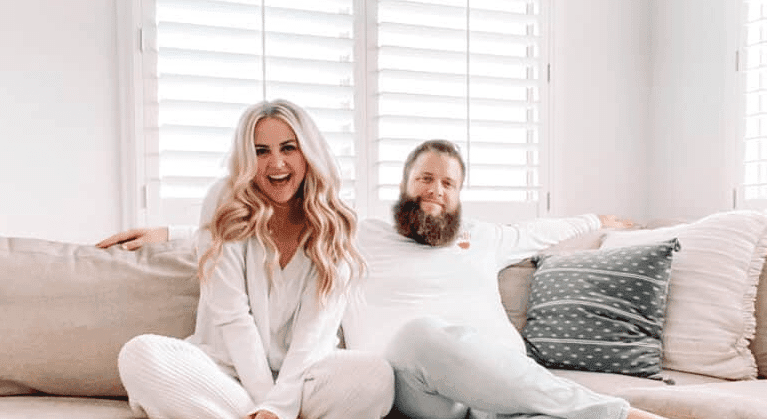A man and woman, Cassie Scroggins, sit together on a couch in front of a window, enjoying each other's company.