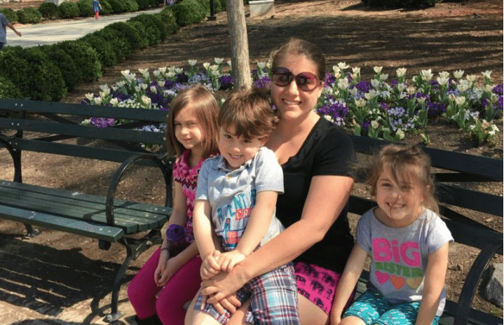 A woman, Lena from What Mommy Does, and three children are seated on a park bench, sharing a joyful moment outdoors.
