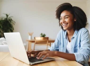 A smiling woman sits at a table with her laptop, engaged in a conversation, radiating warmth and friendliness.