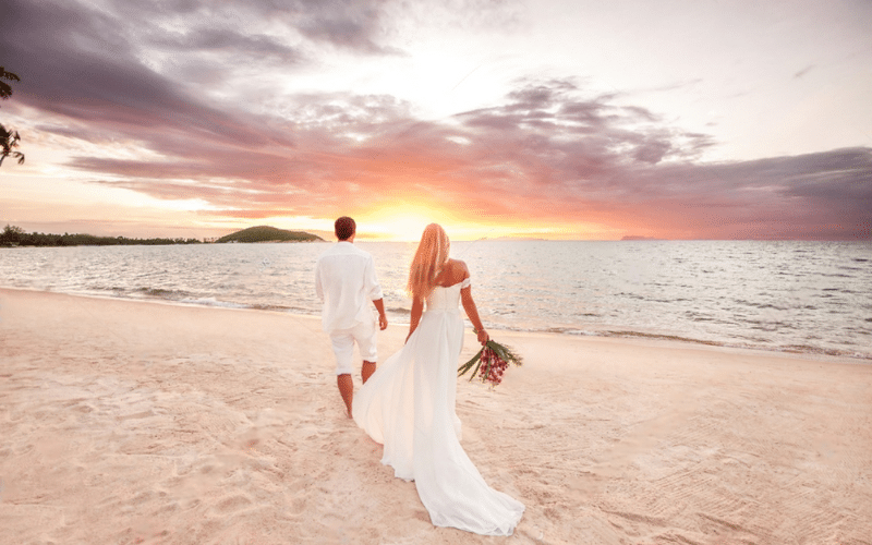 A bride and groom stroll hand in hand along the beach at sunset, showcasing a romantic beach wedding atmosphere.