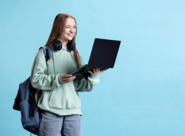 A girl in a green sweater and headphones holds a laptop, focused on making money fast as a teen, ages 12-16.