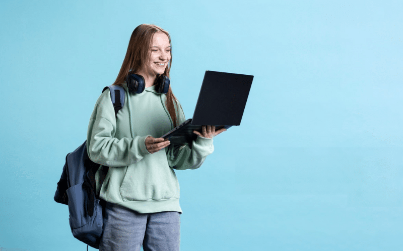 A girl in a green sweater and headphones holds a laptop, focused on making money fast as a teen, ages 12-16.