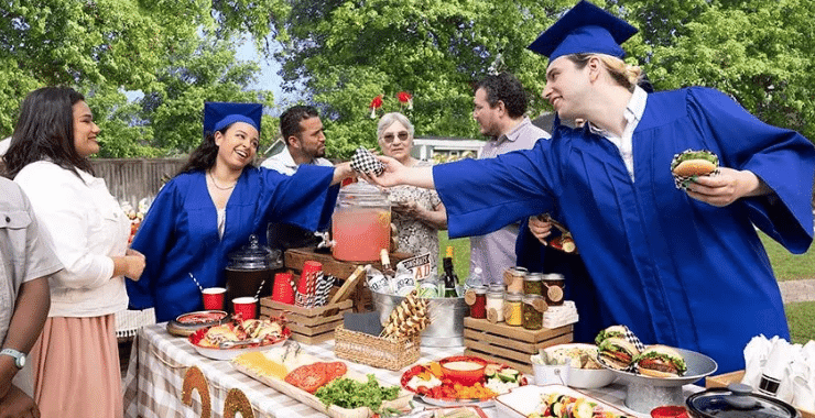 A group of graduates in gowns gathered around a picnic table, enjoying a BBQ celebration after their graduation ceremony.