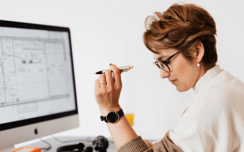 A woman holding a pen while sitting at a desk in an indoor office setting, surrounded by office supplies and a computer. She is wearing clothing appropriate for a professional environment.