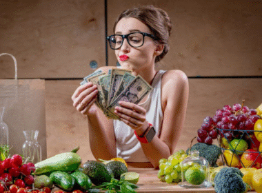 A woman in glasses holds up cash, surrounded by an array of colorful fruits and vegetables on a table.