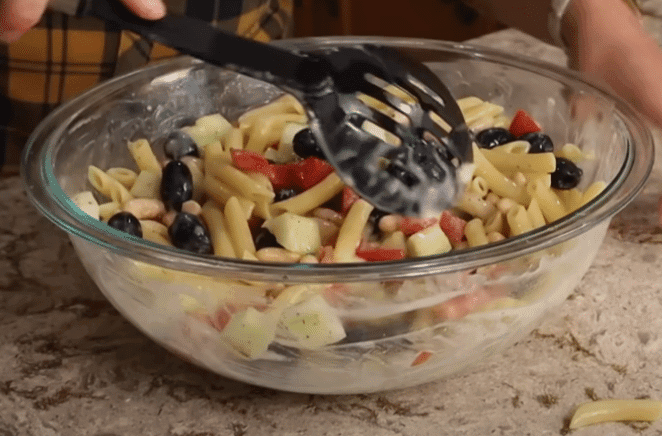 A person mixes colorful pasta salad in a bowl with a large spoon, surrounded by fresh vegetables.