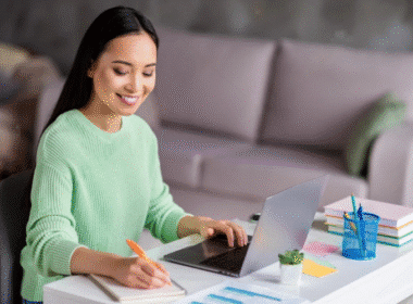 A woman sitting at a desk, focused on her laptop, with a notepad and pen beside her.