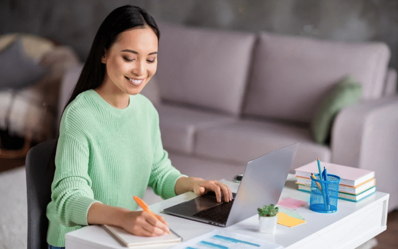 A woman sitting at a desk, focused on her laptop, with a notepad and pen beside her.
