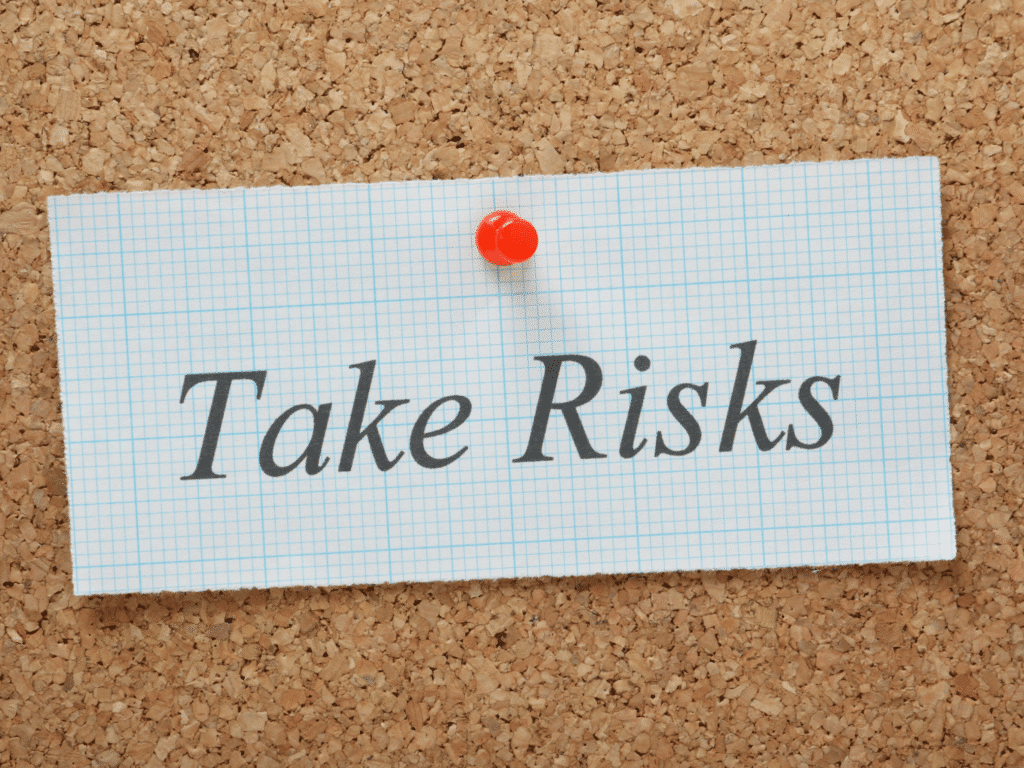 A piece of paper pinned to a cork board with the handwritten text "Take Risks." The paper is rectangular in shape and features a thumbtack holding it in place against the cork surface.