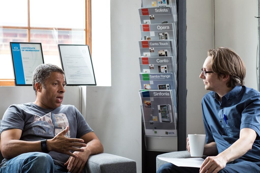 A couple of men sitting in chairs, engaged in conversation indoors. They are surrounded by furniture and have a computer nearby.