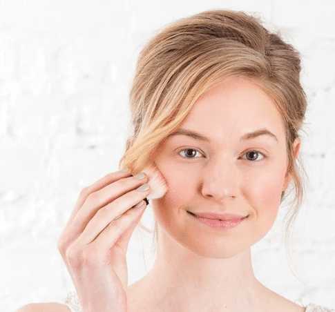 A woman applying minimal makeup with a brush, focusing on her cheeks and eyes in a well-lit setting.