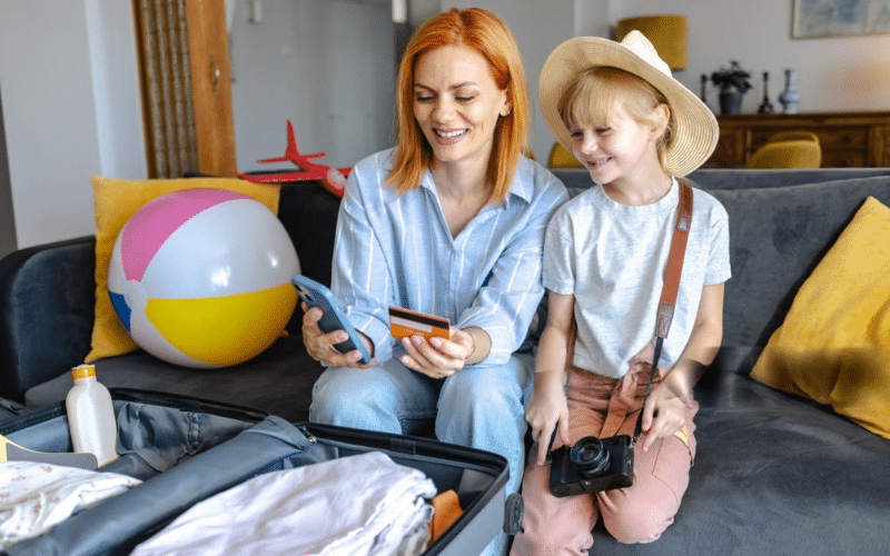 A woman and a child sit on a couch beside a suitcase, preparing for a Vacation Packing List Beach trip together.