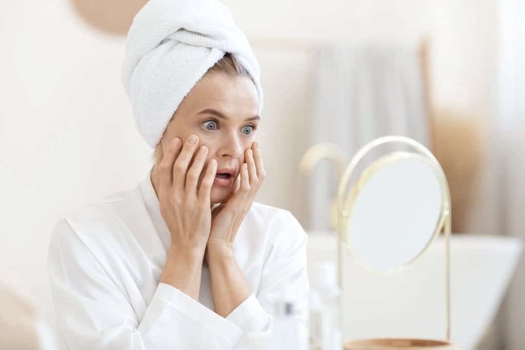 A woman wearing a bathrobe gazes at her face in the mirror, reflecting a moment of self-care.