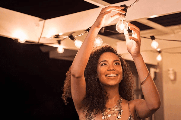 A person adjusts a hanging light bulb amidst glowing string lights, set in a warm, inviting outdoor environment at night.