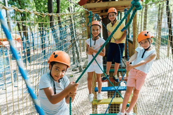 Children navigating a ropes course in a playground, guided by a camp counselor.