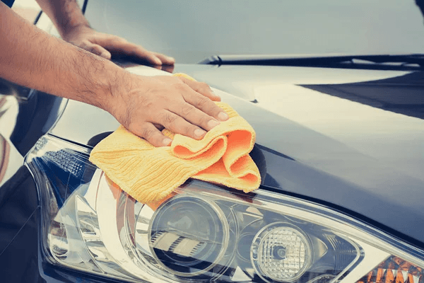 A person wipes the hood of a black car with an orange microfiber cloth, focusing on its shiny surface and headlights.