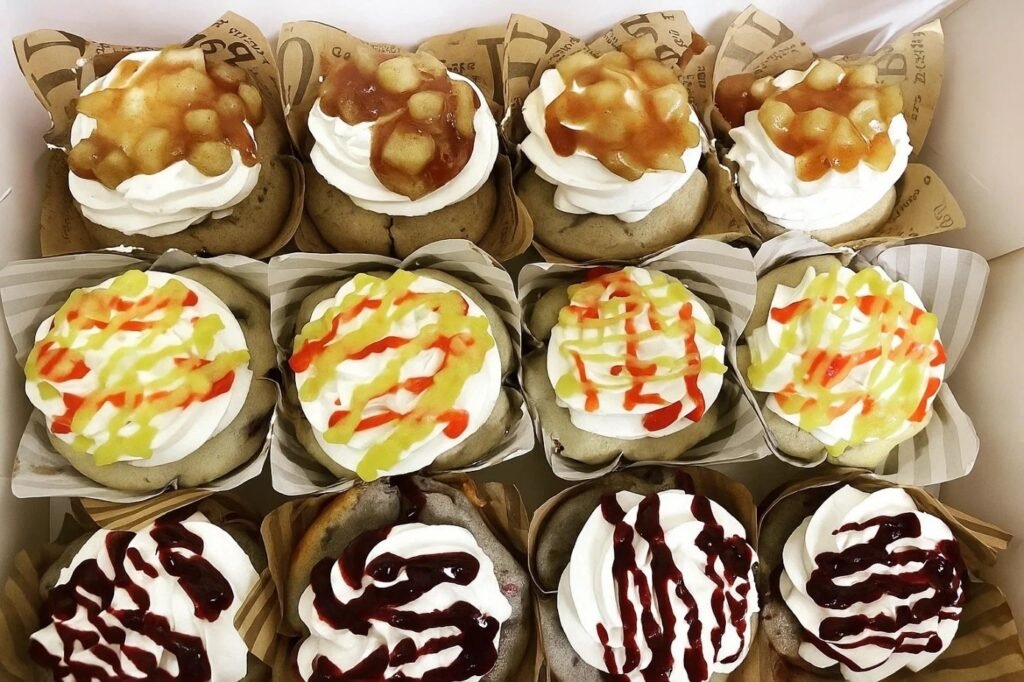 A box of colorful cupcakes topped with various sprinkles and whipped cream, arranged neatly for display.