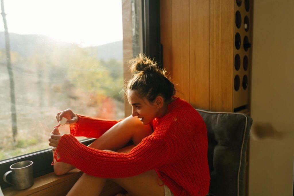 A woman sits on a window sill, enjoying a cup of coffee while contemplating doing hair or nails from home.