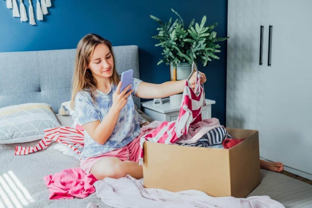 A woman is seated on a bed, with clothes scattered around and a box beside her, suggesting she is sorting through belongings.