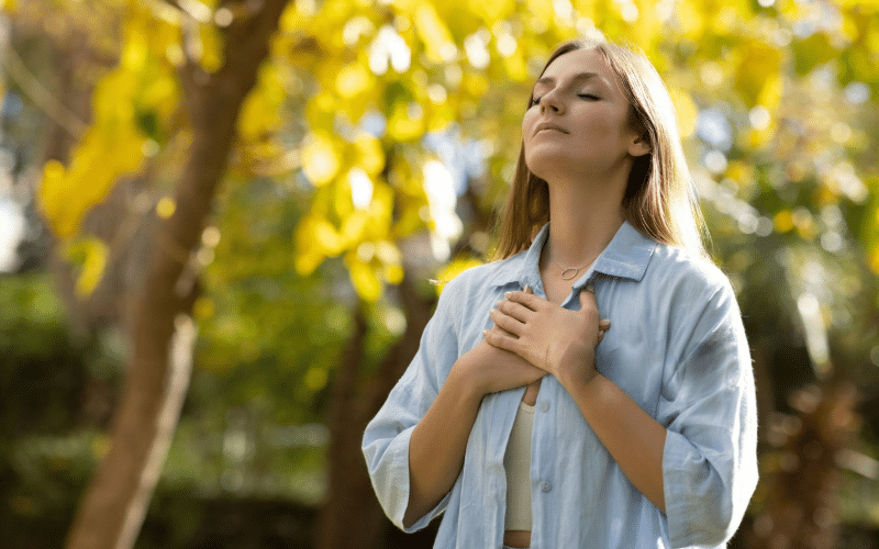 A woman stands before a tree, hands on her chest, reflecting on using prayer to seek employment opportunities.