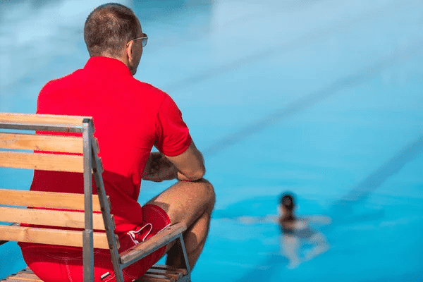 A lifeguard in a red shirt sits on a chair beside a swimming pool, overseeing the area.