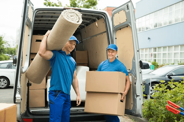 Two workers in blue uniforms loading a rolled carpet and boxes into the back of a moving van, surrounded by parked vehicles and greenery.