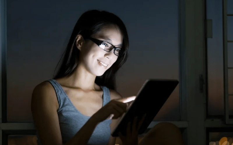 A woman wearing glasses uses a tablet computer at night, focused on her remote night job tasks.