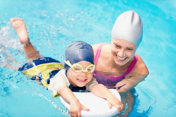 A swim instructor assists a child in a pool, both wearing swim caps, enjoying a lesson together.
