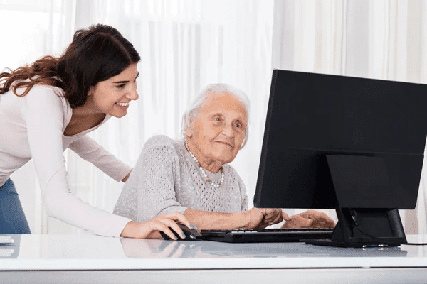 A young woman helps an elderly woman at a computer desk, both focused on the screen, surrounded by bright natural light.