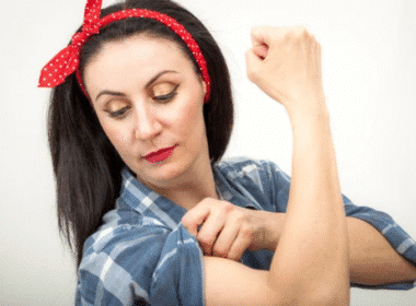 Woman in a blue plaid shirt and red polka dot headband flexes her arm, pulling up her sleeve and looking at her bicep—embodying the spirit of under the table jobs for very hard working adults against a plain background.
