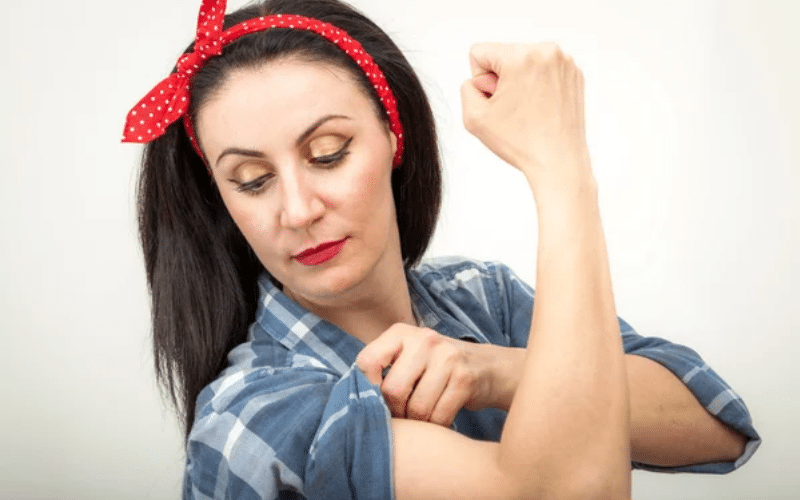 Woman in a blue plaid shirt and red polka dot headband flexes her arm, pulling up her sleeve and looking at her bicep—embodying the spirit of under the table jobs for very hard working adults against a plain background.
