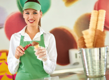 A woman in an apron smiles while holding a waffle cone, showcasing her summer job in a vibrant ice cream shop.