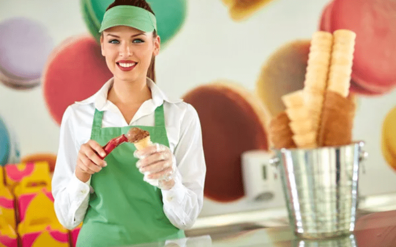 A woman in an apron smiles while holding a waffle cone, showcasing her summer job in a vibrant ice cream shop.