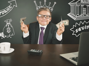 A man in a suit sits at a desk with money and a laptop, illustrating a lesson on teaching kids about money management.