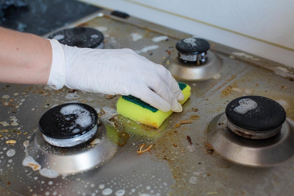 A person wearing gloves is cleaning a stove in an indoor kitchen, surrounded by cookware and bakeware.