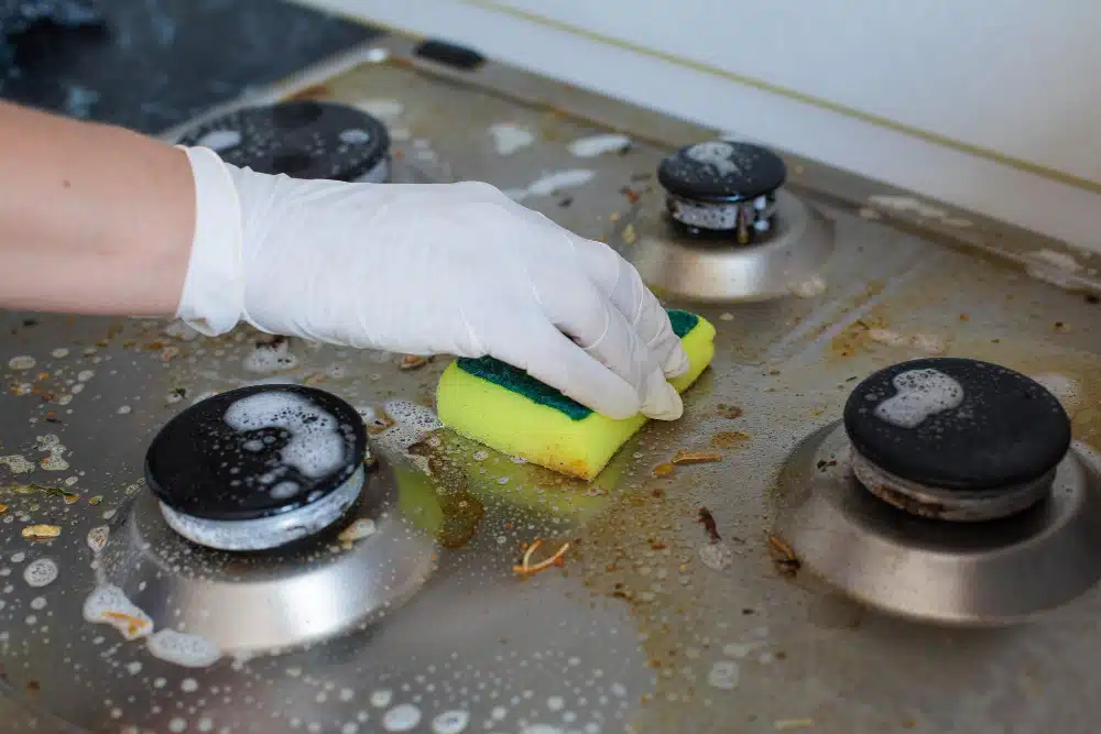A person wearing gloves is cleaning a stove in an indoor kitchen, surrounded by cookware and bakeware.