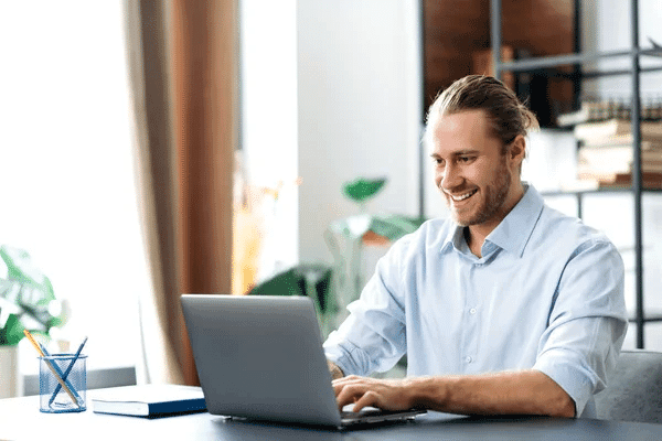A smiling man using a laptop, representing a virtual assistant in a professional setting.