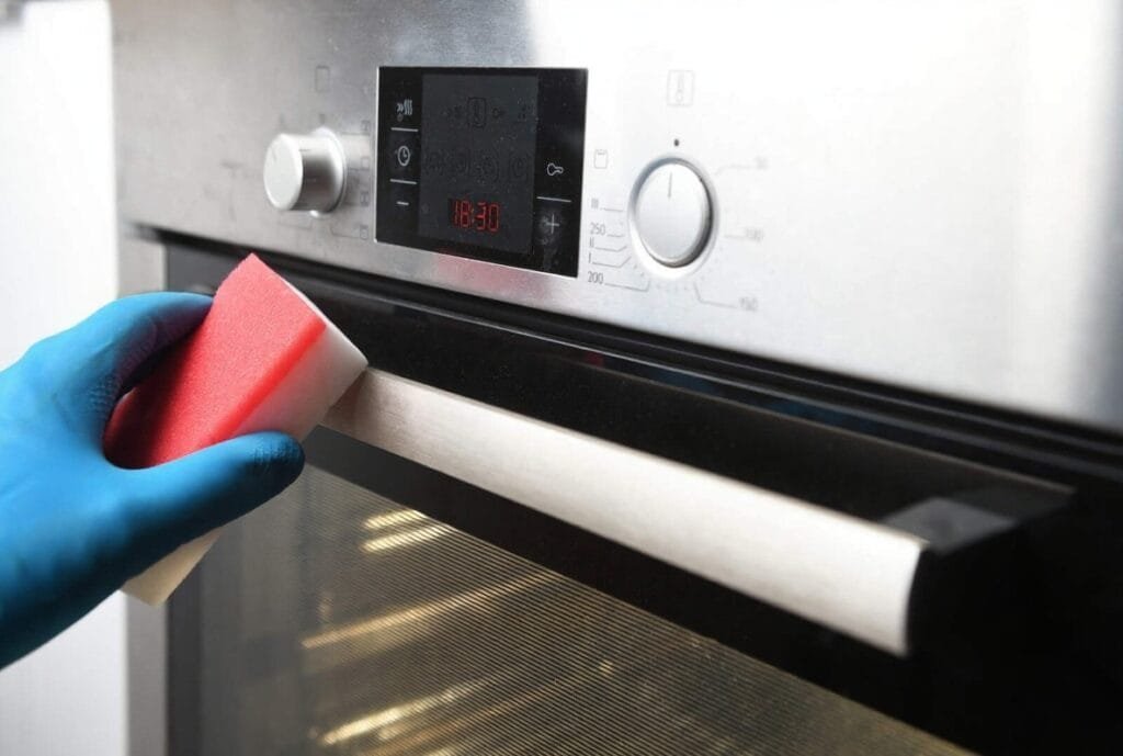 A person wearing blue gloves cleans an oven using white vinegar or Bar Keepers Friend Soft Cleanser.