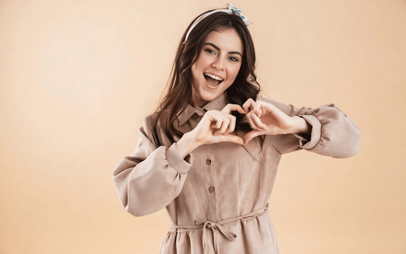 A woman wearing a dress creates a heart sign with her hands, expressing affection and joy.