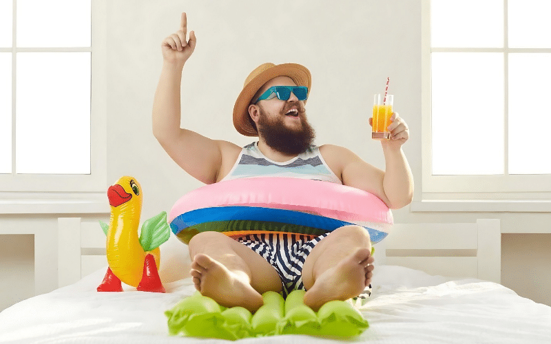 A man with a beard and hat sits on a bed, holding an inflatable ring, suggesting fun birthday staycation ideas.
