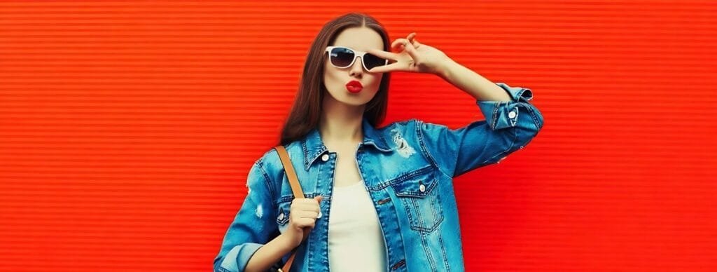 A stylish woman wearing sunglasses and a denim jacket stands against an orange wall, embodying trendy fashion rules for women.