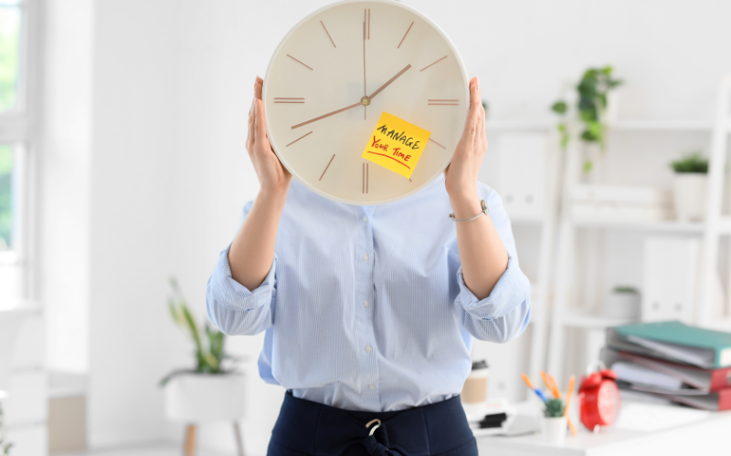 Woman holding a wall clock covering her face with a sticky note that says “Manage Your Time” in a bright office workspace.