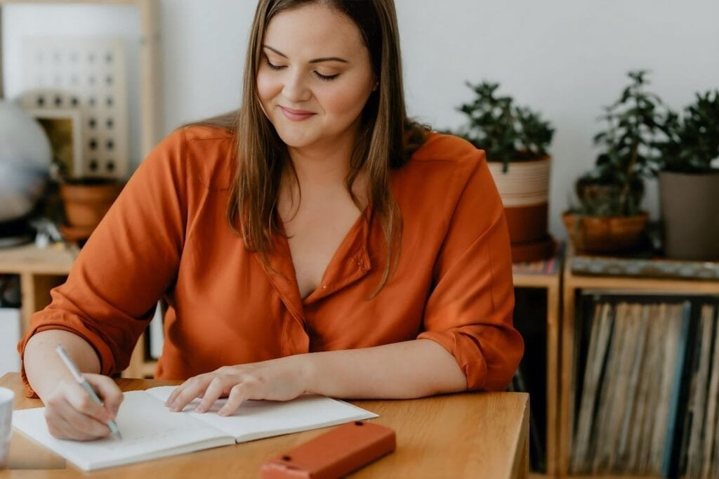 A woman seated at a table writes in a notebook, focusing on her wellness reset reminder.