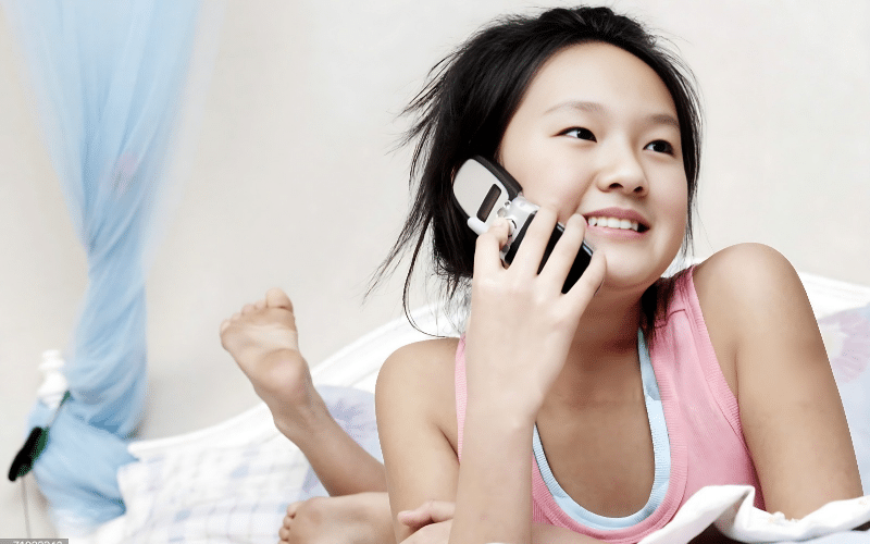 A young girl lying on her bed, chatting on a flip cell phones with a relaxed expression.