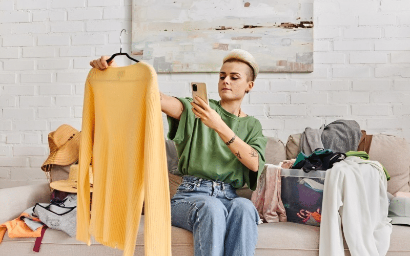 A woman sitting on a couch surrounded by clothes, holding a phone, contemplating how to sell her clothing items.