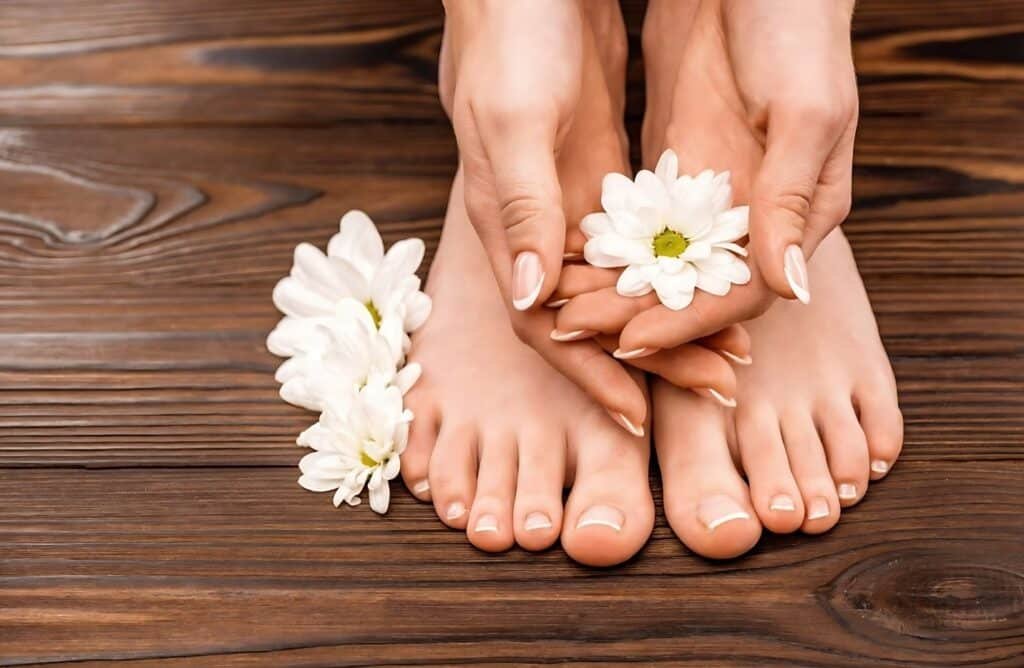 A close-up of a person's hands and feet resting on a wooden floor, adorned with delicate white flowers. The individual is barefoot, showcasing neatly manicured toenails.