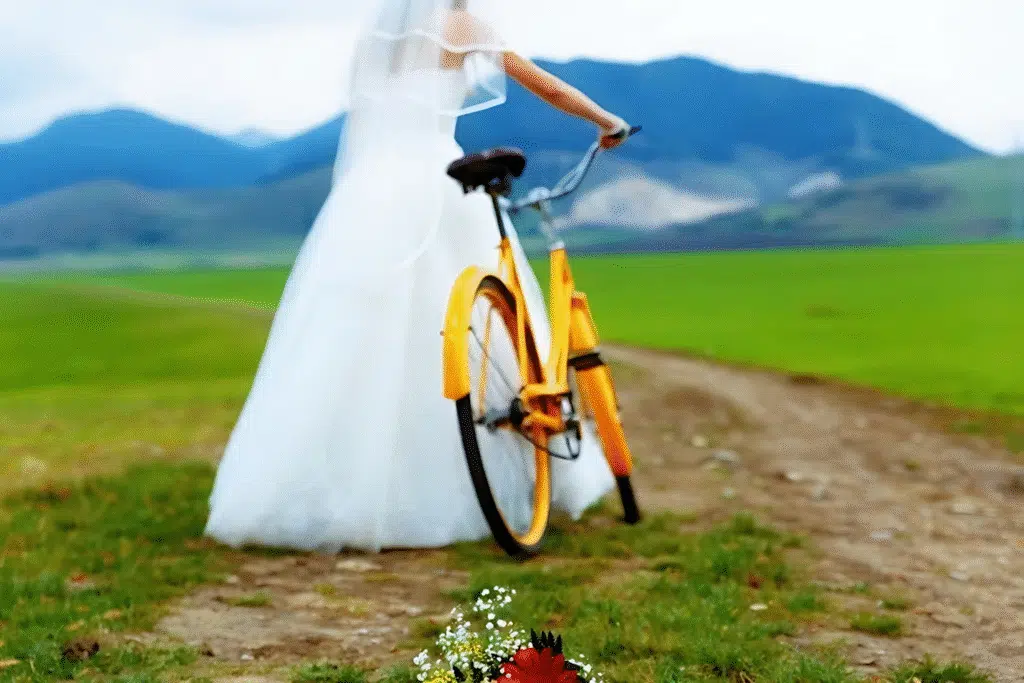 A bride in a white dress stands beside her bicycle in a lush green field, symbolizing a unique wedding moment.